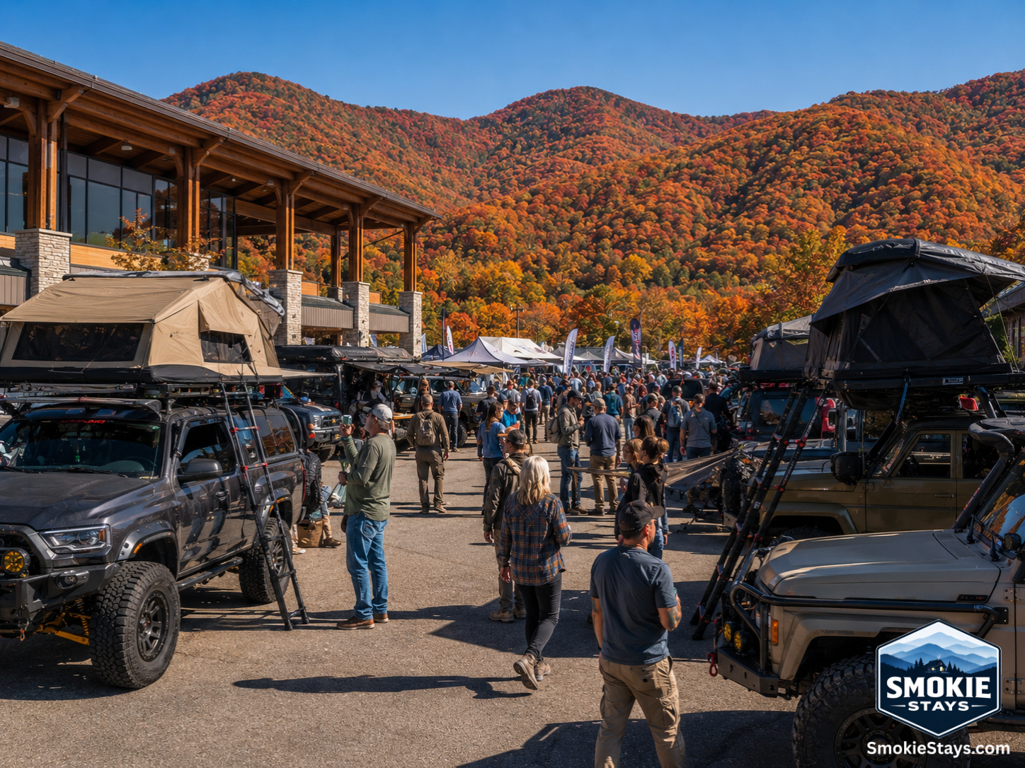 Lifted overland trucks and SUVs with rooftop tents and off-road gear are displayed at the first annual Smoky Mountain Overland Invasion expo at the LeConte Center in Pigeon Forge, with fall foliage on the Smoky Mountain ridgelines in the background.